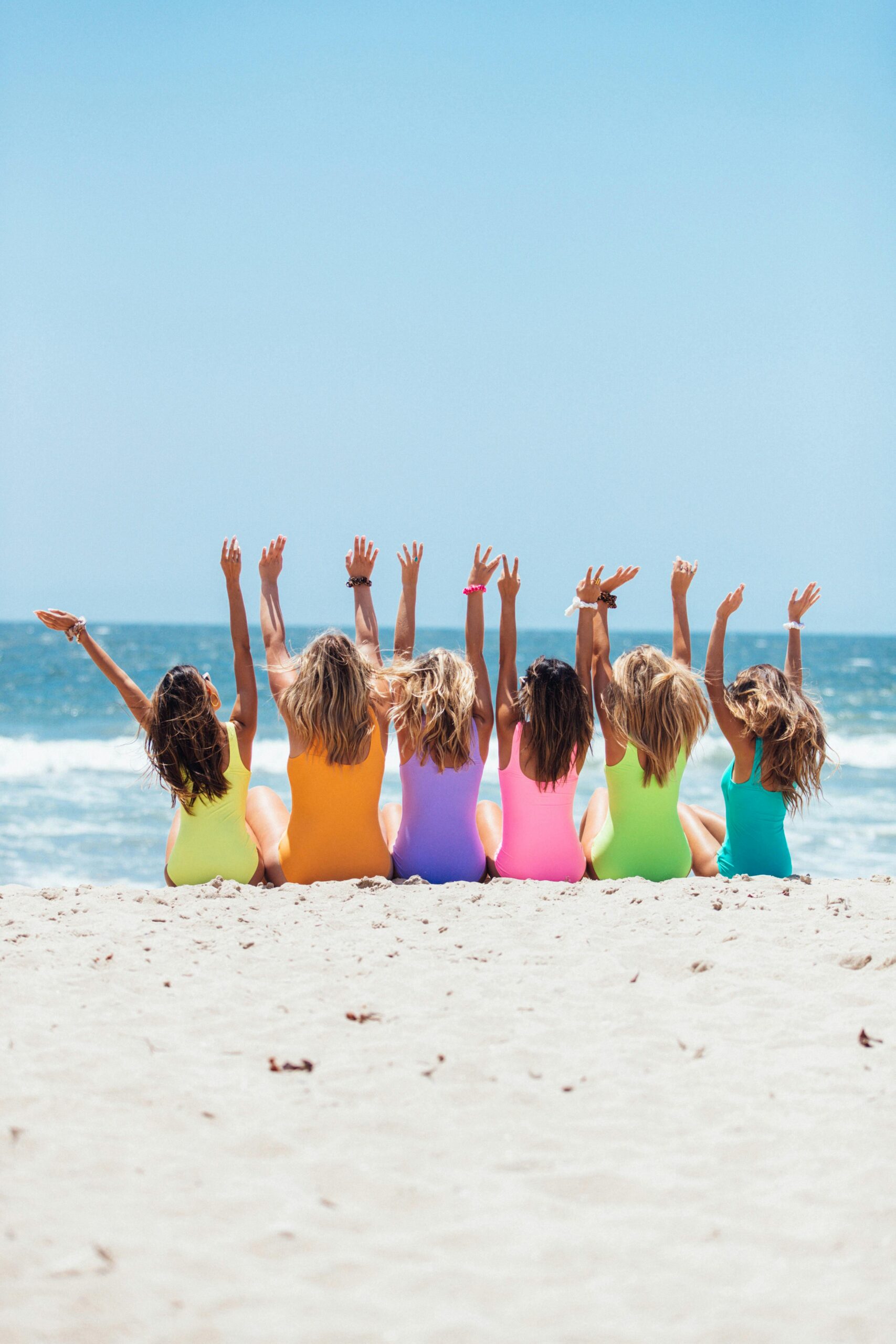 Group of women in colorful swimsuits sitting on a beach raising their arms toward the sea — symbol of women supporting women on their fertility journey