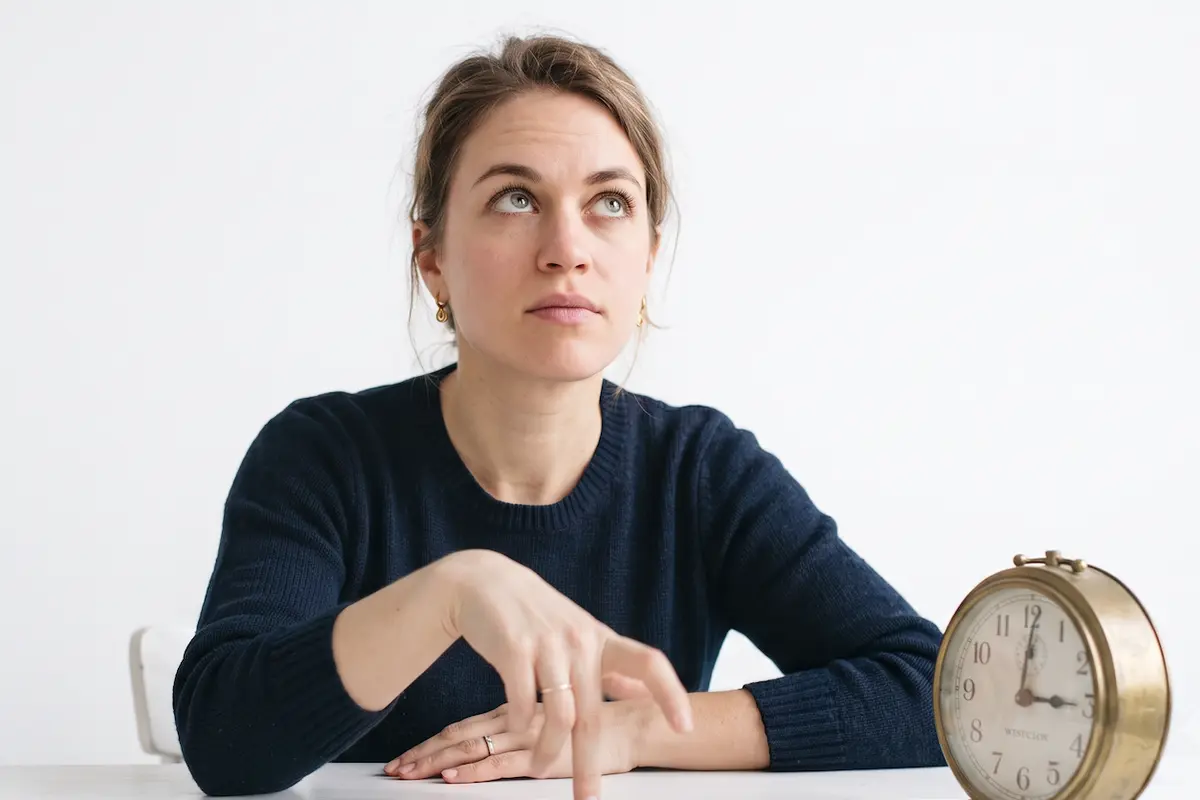 A woman at a table drumming her fingers beside an alarm clock, looking up — the long two-week wait after IVF embryo transfer