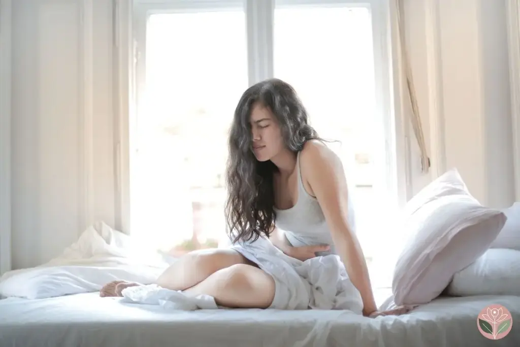 Woman sitting on a white bed by a bright window, holding her lower abdomen in pain — endometriosis and fertility