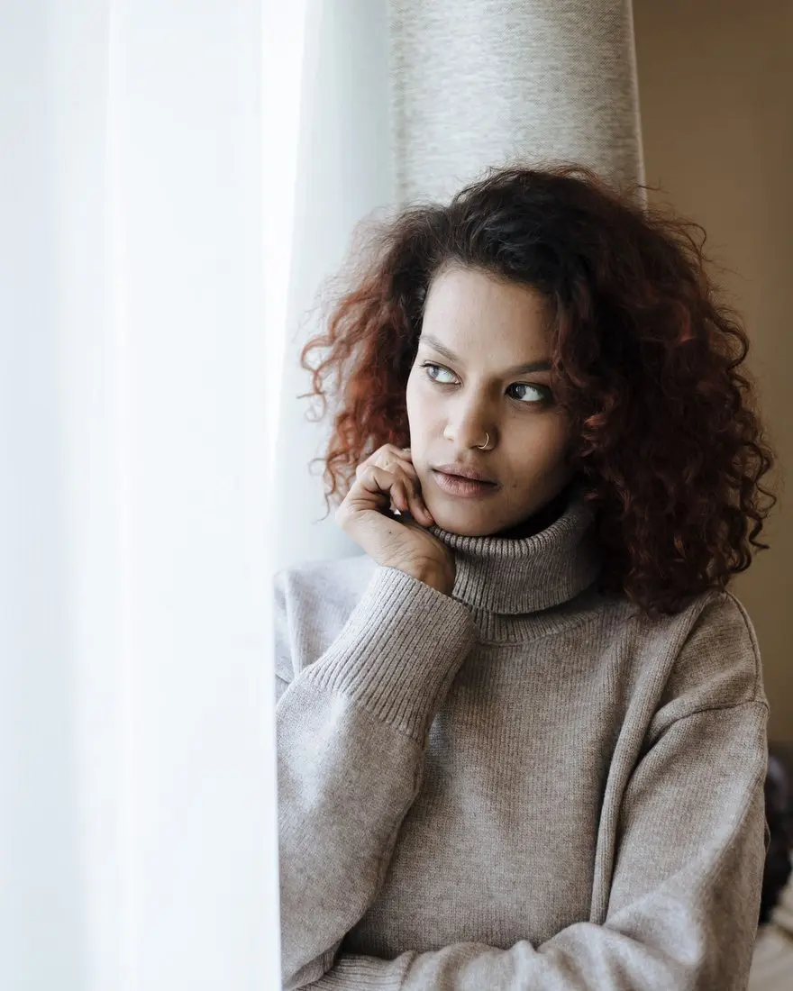 A woman in a soft grey jumper standing by a bright window, lost in thought — making sense of an endometriosis diagnosis